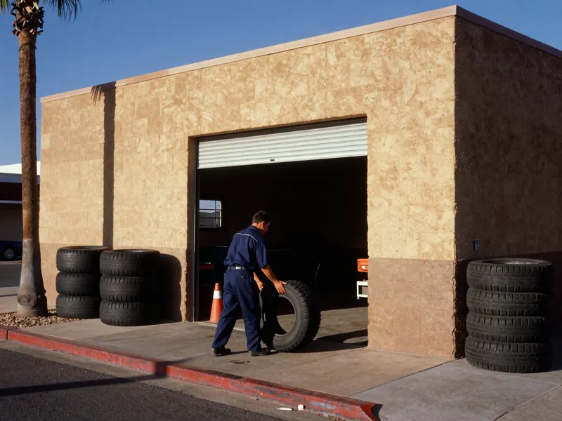 A mechanic in navy coveralls rolls a tire into the open roll-up bay of a tan block shop in low desert sun, with stacked tires on both sides, a palm tree at left and a red-painted curb in front.